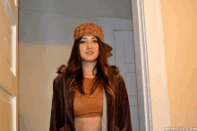 a woman wearing a hat and a brown jacket is standing in a doorway with memeful.com in the corner