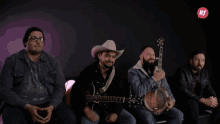 a group of men are sitting on a couch playing guitars and banjos with a rf logo in the background