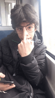 a boy wearing glasses and a black jacket holds his middle finger to his nose