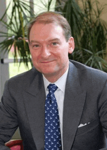 a man wearing a suit and tie is sitting in front of a plant .