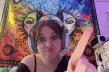 a woman wearing headphones is giving the middle finger in front of a tapestry with the sun and moon on it