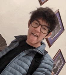 a young man wearing glasses and a blue jacket sticks out his tongue