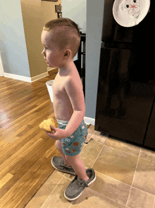 a shirtless toddler standing in front of a black refrigerator with a paper plate on it