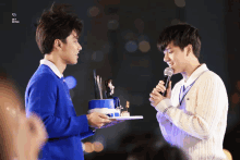 a man holding a microphone stands next to another man holding a blue cake