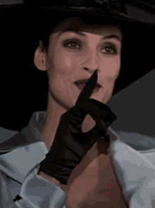 a woman wearing a hat and a black glove holds her finger to her mouth