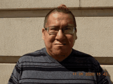 a man wearing glasses and a striped shirt is standing in front of a wall and looking at the camera .