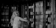 a black and white photo of a woman standing in front of a glass cabinet filled with plates and cups .