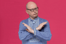 a bald man wearing glasses and a blue shirt is crossing his arms in front of a pink background