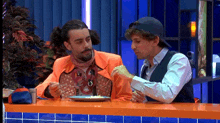 two men are sitting at a table with a plate of food in front of them