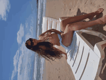 a woman in a bikini sits on a white chair on the beach