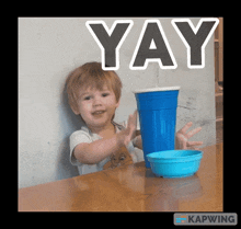 a little boy is sitting at a table with a blue cup and a blue bowl and the word yay above him