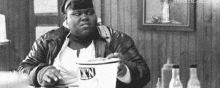 a black and white photo of a fat man sitting at a table holding a bucket of ice cream .