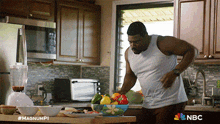 a man in a white tank top is standing in a kitchen with a nbc logo on the bottom right