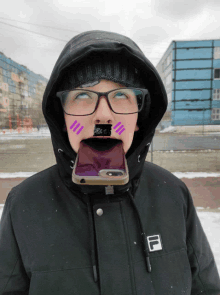 a person wearing glasses and a fila jacket holds a cell phone in their mouth