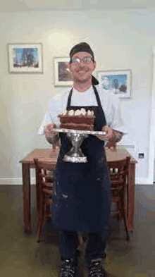 a man in an apron is standing in front of a table holding a cake on a plate .