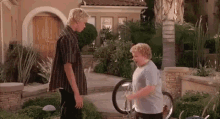 two boys are standing next to each other in front of a house while one of them is holding a bicycle wheel .