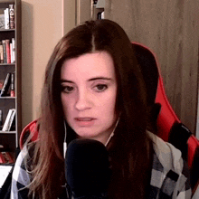 a woman wearing headphones and a plaid shirt is sitting in front of a microphone in front of a bookshelf .