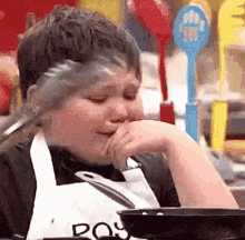 a young boy is sitting at a table with a pan in front of him and a spoon in his mouth .