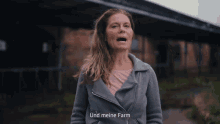 a woman in a gray jacket stands in front of a building with the words " und meine farm " on the bottom