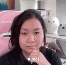 a woman is sitting in front of a pink chair with her hand on her chin and the word kudos written on her face .