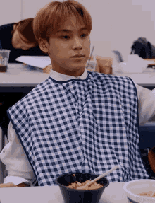 a young man wearing a blue and white checkered apron is sitting at a table with a bowl of food and chopsticks .