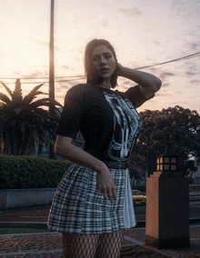 a woman wearing a black t-shirt and a plaid skirt stands on a sidewalk