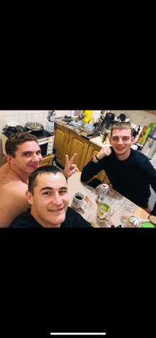 three men are sitting at a table in a kitchen and one is giving a peace sign