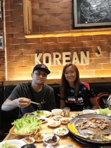 a man and a woman are sitting at a table in front of a wall that says korean