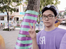 a young man wearing glasses and a purple shirt is standing next to a tree with purple and green tape on it .