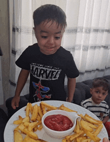 a boy wearing a shirt that says marvel stands next to a plate of french fries