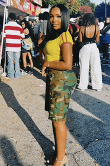 a woman wearing a yellow shirt and camo shorts stands in front of a crowd of people