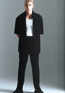 a man wearing a black shirt and black pants stands in front of a white wall