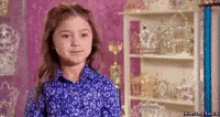 a little girl in a blue shirt is standing in front of a shelf with a lot of decorations on it .