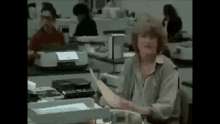 a woman is sitting at a desk in an office holding a piece of paper and talking on a phone .