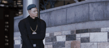 a man wearing a turtleneck and a gold chain is standing in front of a brick wall