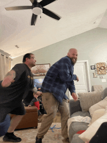 two men are dancing in a living room with a ceiling fan behind them