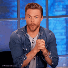 a man with a beard and a denim shirt is sitting at a table with his hands folded in front of a window .