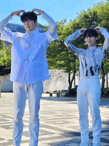 two young men in white clothes are standing next to each other making a heart shape with their hands