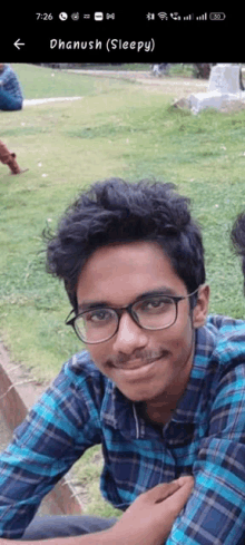 a young man wearing glasses and a plaid shirt is sitting in the grass and smiling for the camera