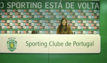 a woman sits at a desk in front of a wall that says o sporting esta de volta