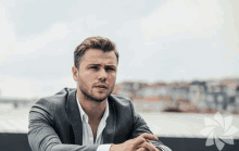 a man in a suit and white shirt is sitting with his hands folded in front of a body of water