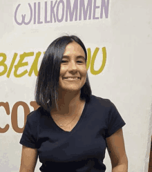 a woman in a black shirt smiles in front of a sign that says willkommen