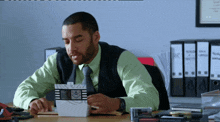 a man in a green shirt and tie sits at a desk in front of binders with the letter a on them