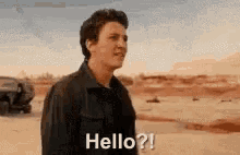 a man in a black jacket is standing in the desert and says `` hello '' .