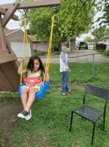 a little girl is sitting on a swing while a boy stands behind her .