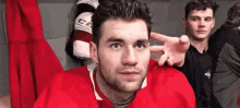 a man in a red hockey jersey is sitting in a locker room making a peace sign with his hands .