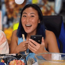 a woman is sitting at a table with chopsticks and a bowl of food and holding a cell phone .