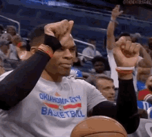 a man wearing an oklahoma city basketball shirt is sitting in a stadium with his hands in the air .