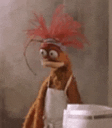 a muppet wearing an apron and a red feathered hat is standing next to a blender .