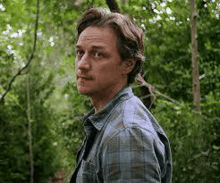 a man in a plaid shirt is standing in the woods looking to the side .